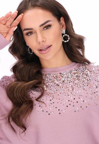 Világos rózsaszín bordázott pulóver, amelyen szórt flitter díszítések találhatók az elején. A modellen ezüst karika fülbevalók vannak, és hullámos haja van.