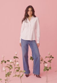 Frau mit langen dunklen Haaren, die ein gestreiftes weißes Hemd, blaue Jeans und schwarze Absätze trägt, steht zwischen in Vasen arrangierten rosa Rosen vor einem pinken Hintergrund.