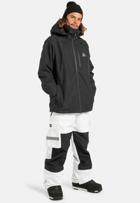 Czarna wodoodporna kurtka z wysokim kołnierzem i kieszenią na zamek z przodu, zestawiona z białymi i czarnymi spodniami cargo z dużymi kieszeniami i paskami.