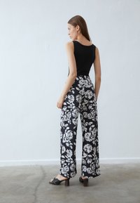 Haut sans manches noir associé à un pantalon large avec un motif floral blanc. La mannequin porte des chaussures à talons noirs ouvertes. Textures lisses.