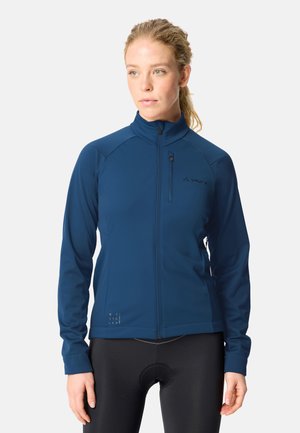 Vrouw draagt een blauwe jas met rits en een borstzakje en zwarte fietskleding, staand tegen een effen lichte achtergrond.