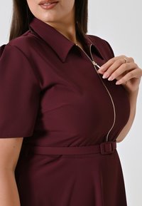 Eine Frau trägt ein kastanienbraunes Kleid mit einem Reißverschluss vorne und einem passenden Gürtel, während sie den Reißverschlussgriff nahe dem Kragen hält.