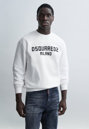 Sudadera blanca con la inscripción "DSQUARED2 MILANO" en negro, combinada con vaqueros oscuros desgastados. El modelo posa frente a un fondo claro.