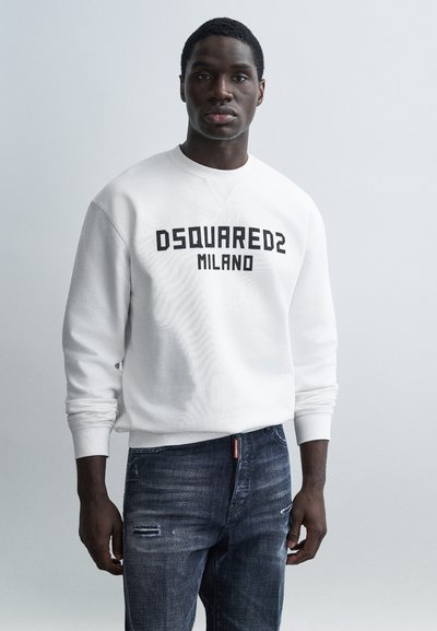 Sweat-shirt blanc avec le texte "DSQUARED2 MILANO" en noir, porté avec un jean en denim sombre déchiré. Le mannequin pose devant un fond clair.
