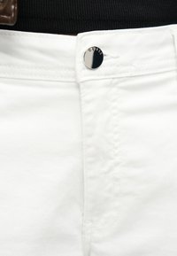 Κοντινό πλάνο λευκών τζιν τζην με ασημί κουμπί χαραγμένο με τη λέξη "GUESS", φορεμένα με μαύρο ριμπ μπλουζάκι.