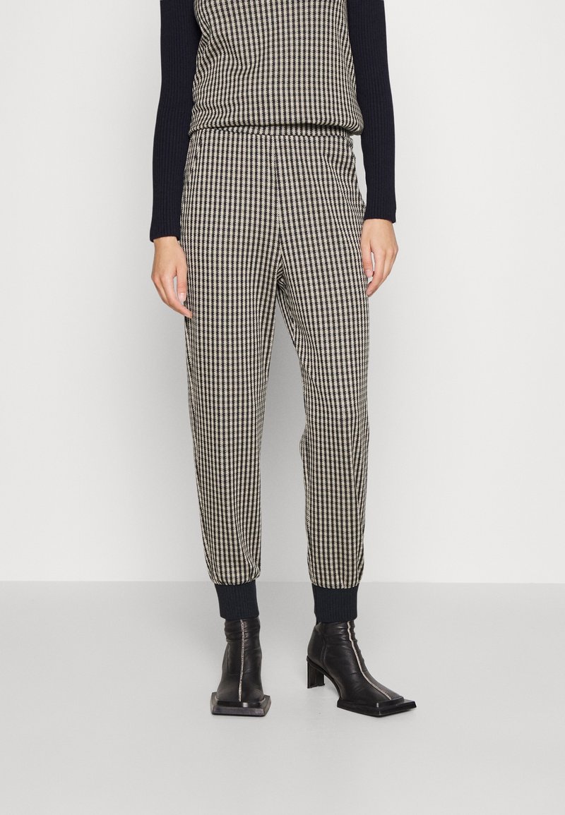 Un pantalon à carreaux présente un motif de grille noir et crème, avec une coupe décontractée, une taille élastique et des poignets côtelés. Associé à des bottines noires.