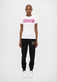 Hvitt kortermet t-skjorte med fet rosa "VERSACE JEANS COUTURE"-tekst, kombinert med sorte sweatpants med en gull logoakcent.