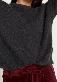 Pull en maille gris foncé avec une texture côtelée, col rond et coupe décontractée, associé à un pantalon en velours côtelé bordeaux avec ceinture à nouer.