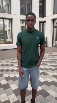 Polo verde con colletto ed emblema, abbinato a pantaloni corti in denim azzurro chiaro. Il modello è in piedi su uno sfondo di pavimentazione a motivo.