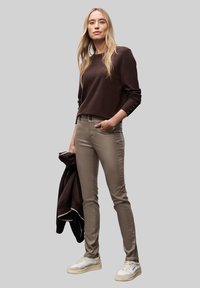Top de manga larga marrón, pantalones ajustados color beige claro y zapatillas blancas. Textura de tela suave; el top tiene puños abotonados. Chaqueta colgada del brazo.