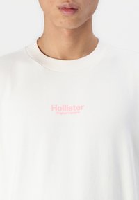 T-shirt en coton blanc avec un col ras du cou, présentant un logo brodé rose qui indique "Hollister Original Designs" sur la poitrine.