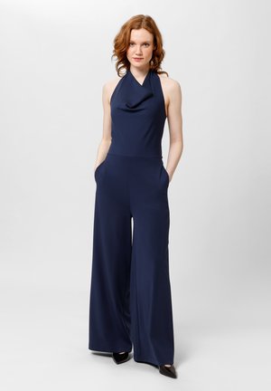 Mono azul marino con cuello drapeado, piernas anchas y bolsillos laterales. Hecho de tejido suave, combinado con tacones negros puntiagudos.