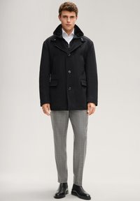 Cappotto nero in lana con collo texturizzato, tasche anteriori e bottoni, abbinato a pantaloni grigi a quadri e stivali neri.