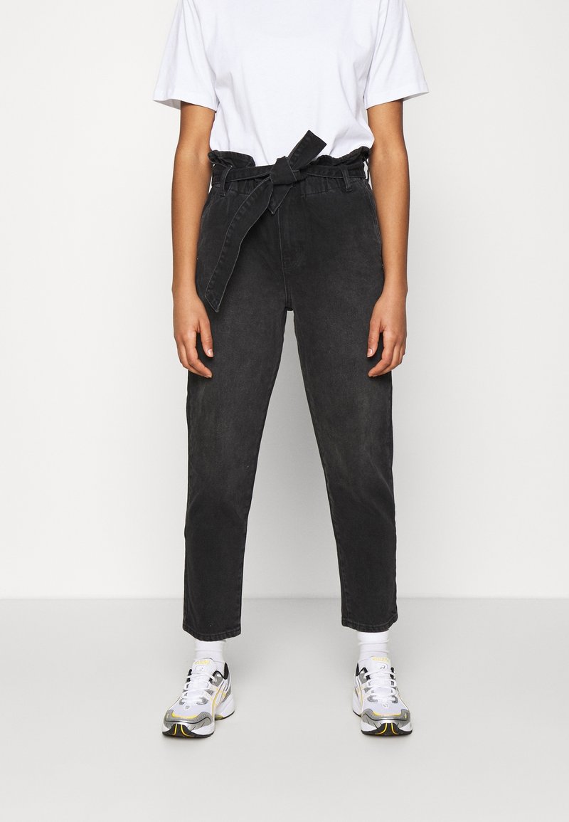 Pantalon en denim noir à taille haute, avec un détail de ceinture à nouer et un design de jambe fuselée, associé à un t-shirt blanc et des chaussures de sport.