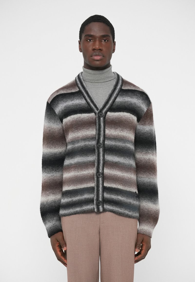 Triibuline kardigan V-kaelusega, millel on halli, pruuni ja musta värvi triibud. Valmistatud pehmest materjalist, sulgub nööpidega ja pikad varrukad.