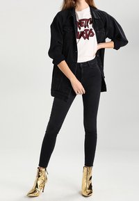 Černá džínová bunda přes bílý grafický tričko, černé skinny džíny a lesklé zlaté kotníkové boty se zipem.