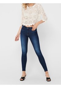 Weißes gestricktes Crop-Top mit gewelltem Saum und floralen Mustern, kombiniert mit dunkelblauen Skinny-Jeans und schwarzen Rutsch-Sandalen.