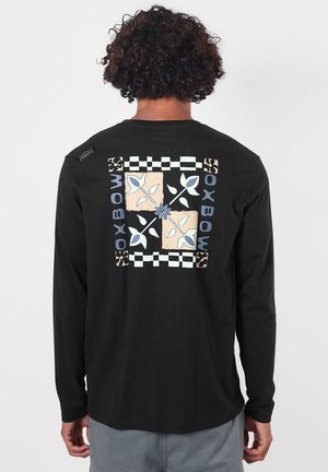 Personne portant un t-shirt noir à manches longues avec un motif de quatre carrés floraux et le mot "OXBOW" en lettres bleues dans le dos.