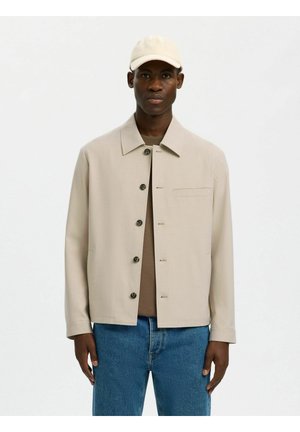 Mann iført beige knappet jakke, brun skjorte, blå jeans og beige baseballcaps mot ensfarget hvit bakgrunn.