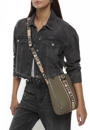 Kobieta w przyciętej czarnej dżinsowej kurtce i dżinsach, nosząca oliwkową torbę na ramię z beżowym paskiem i czarnym napisem.