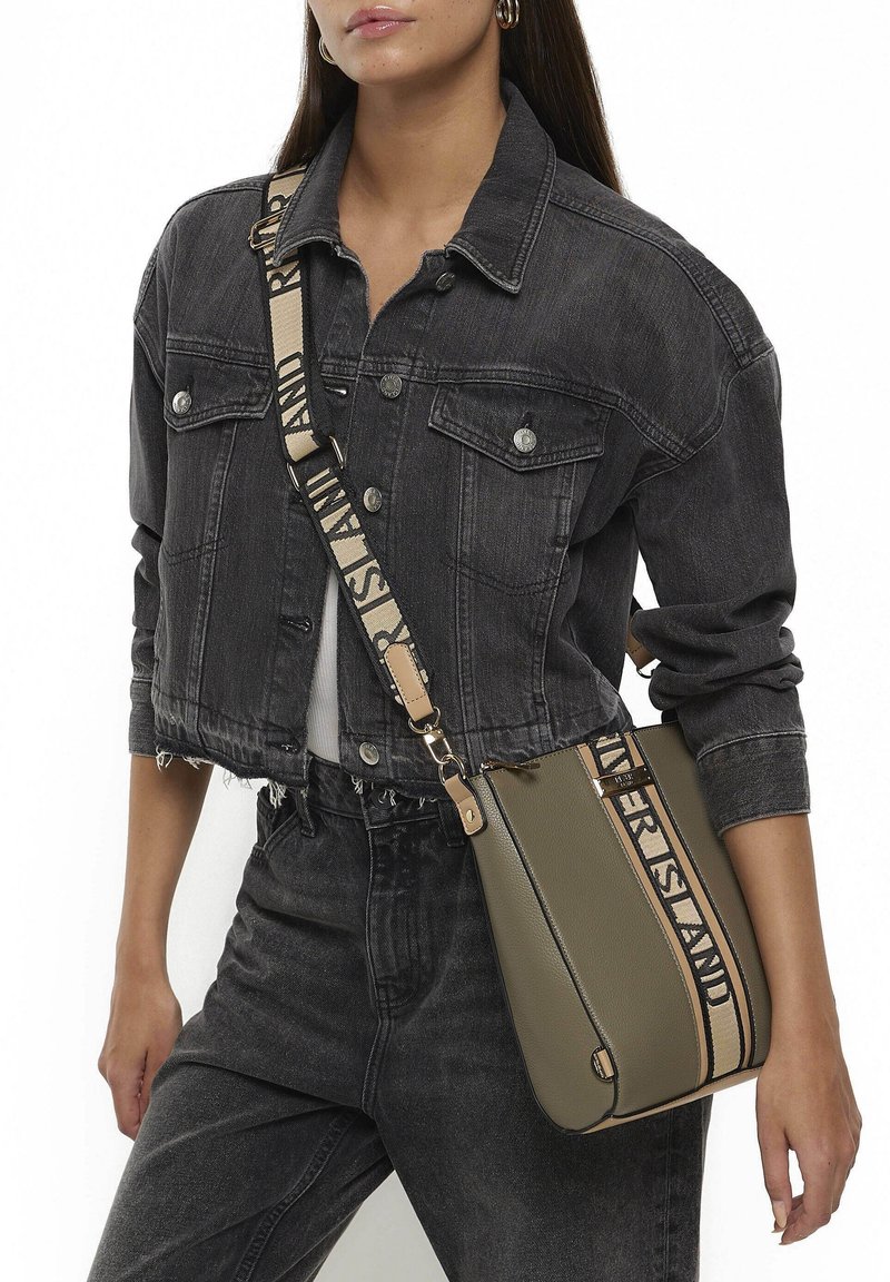 Žena v černé cropped džínové bundě a džínách, nese olivově zelenou kabelku přes rameno s béžovým popruhem a černým potiskem.