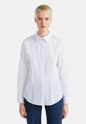 Camicia bianca a maniche lunghe con colletto classico, chiusura a bottoni e orlo curvo, realizzata in tessuto morbido, abbinata a pantaloni in denim blu.