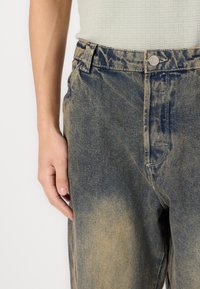 Fakó kék színátmenetes farmernadrág, zipzáras és gombos kivitelben. Az anyag pamutban gazdag, enyhén durva tapintással.