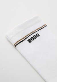 Biely bavlnený ponožka s rebrovanou textúrou, s čierno-zlatým pruhovaným pásom na vrchu a nápisom "BOSS" v čiernej výšivke.