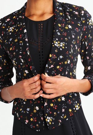 Person iført en sort blazer med flerfarvede små blomsterprint, som lukker fronten over en sort kjole med blonde detaljer.