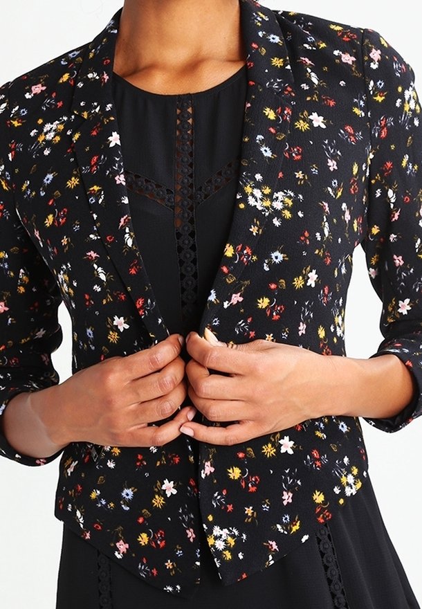 Personne portant un blazer noir avec de petits motifs floraux multicolores, fermant le devant sur une robe noire avec des détails en dentelle.