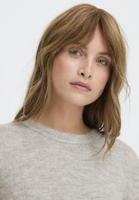 Fiatal nő, világosbarna hullámos hajjal, világosszürke kötött pulóvert visel, enyhén oldalra néz, semleges arckifejezéssel.