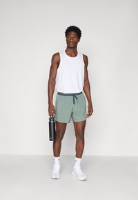 Conjunto atlético masculino que incluye una camiseta sin mangas blanca y pantalones cortos verdes. Sostiene una botella de agua negra. Zapatillas blancas y detalles de tela texturizada.