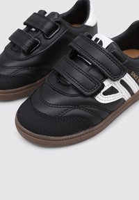 Zapatos deportivos negros con material texturizado negro, detalles en blanco, dos correas de Velcro y una suela de goma marrón. Presentan detalles de costura curvada.