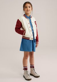Röd och krämfärgad varsityjacka över en denimklänning, parat med beige snörstövlar och rödrandiga strumpor. Enkel skärning och avslappnad design.