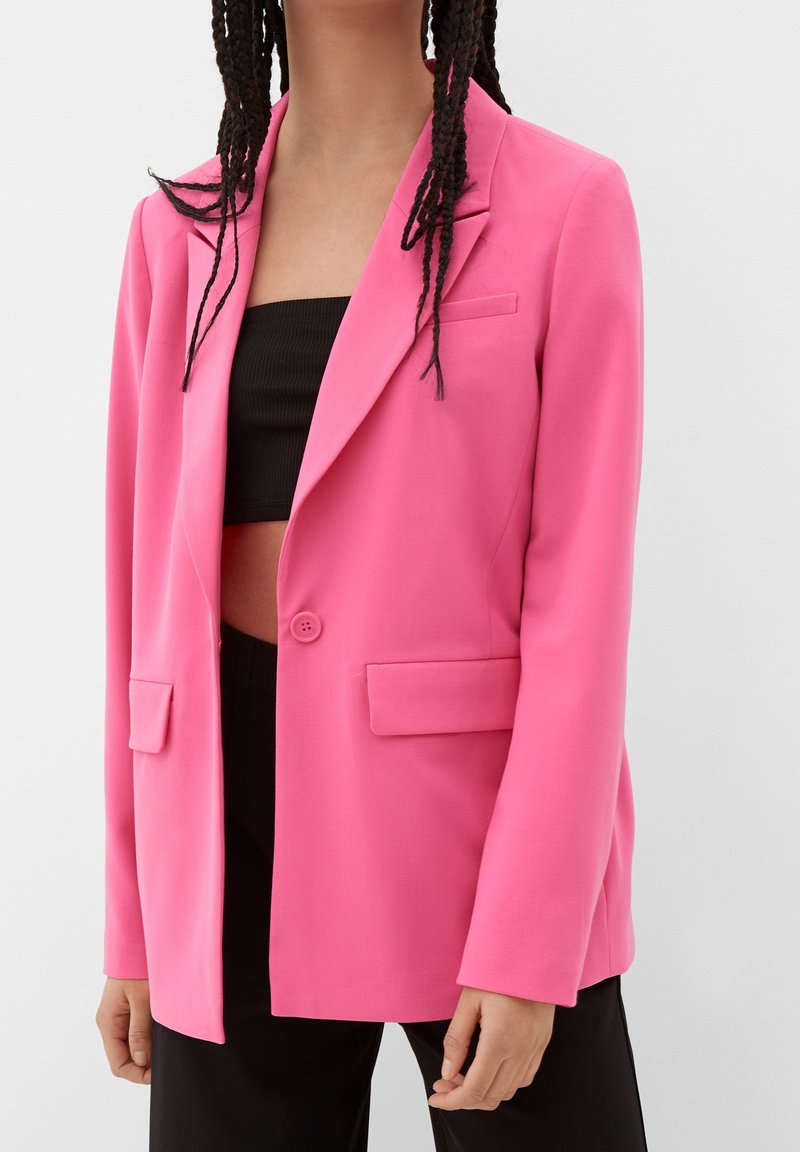 Blazer rose en tissu lisse avec une coupe cintrée, fermeture à un bouton, deux poches avant et revers crantés. Crop top noir en dessous.