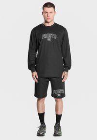 Czarna koszulka z długim rękawem i szorty z białym logo "PROSTO". Osoba nosi czarne skarpetki i kamuflażowe buty. Gładkie tło.