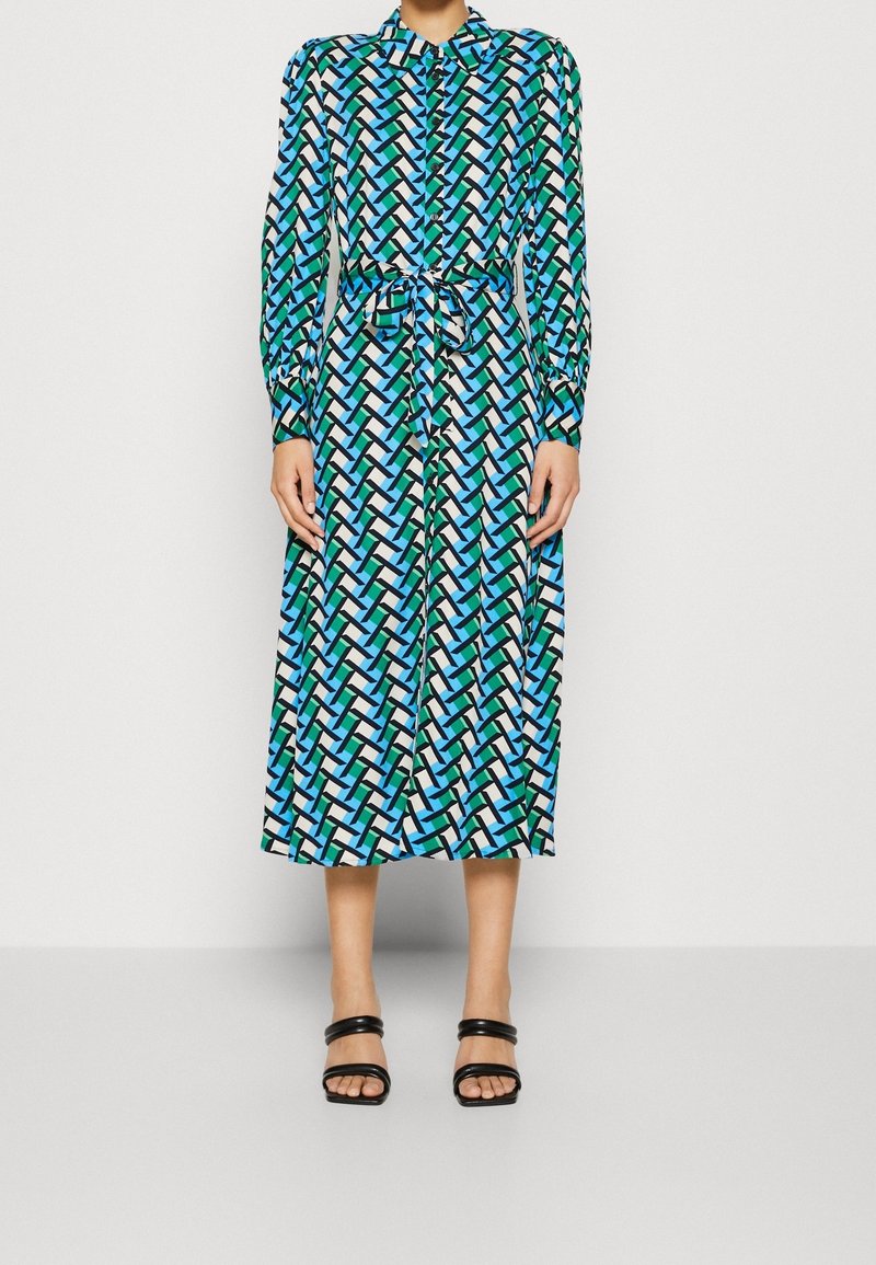 Femme portant une robe midi à manches longues boutonnée avec un imprimé géométrique bleu, vert et noir, ceinture nouée et sandales noires ouvertes.
