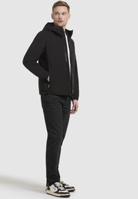 Black soft-shell jacket with a hood, zip closure, elastic cuffs, and side pockets, paired with dark jeans and black-and-white sneakers.