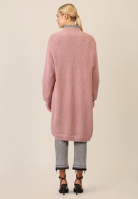 Lange roze gebreide cardigan met een geribbelde textuur, gedragen over grijze cropped jeans met rafelige zoom en zwarte hakken met banden. Eenvoudig ontwerp.