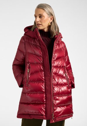 Frau trägt einen glänzenden roten, wattierten Wintermantel mit Kapuze und Reißverschlusstaschen, steht mit den Händen in den Taschen und schaut zur Seite.