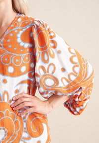 Blusa arancione e bianca con audaci motivi floreali, maniche a sbuffo e scollo a V. Realizzata in tessuto leggero con una finitura testurizzata.