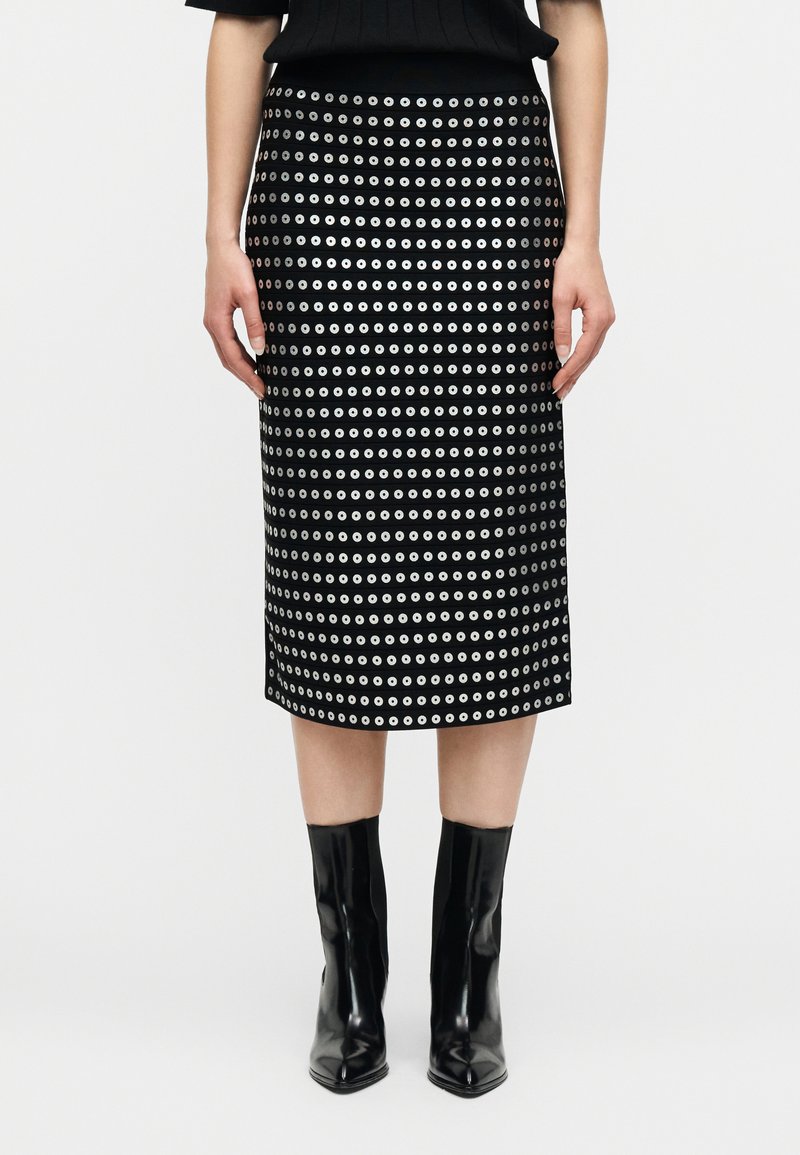 Γυναίκα φοράει μαύρη μίντι φούστα με οριζόντιες σειρές ασημένιων οπών και μαύρα μπότες αστραγάλου με μυτερή μύτη σε λευκό φόντο.