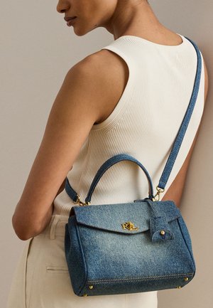 Femme portant un débardeur blanc côtelé et un pantalon crème, portant un sac à main en jean bleu avec des détails dorés sur l'épaule.