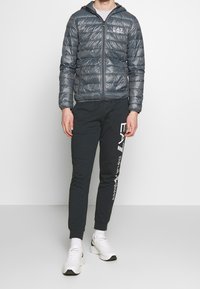 Šedá péřová bunda s prošívanou texturou a kapucí, v kombinaci s tmavými tepláky s bočním logem. Model nosí bílé tenisky.