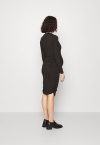 Robe longue à manches longues noire à côtes avec un design froncé à la jupe, assortie à des chaussures à talons blocs noires. Sur fond uni.