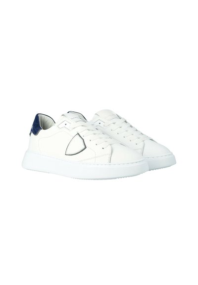 Witte lage sneakers met marineblauwe hieltabs, witte veters en een schildvormige badge aan de zijkant, weergegeven op een witte achtergrond.