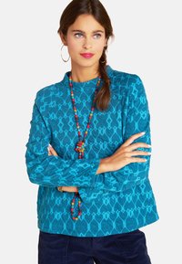 Türkises, langärmeliges Oberteil mit strukturiertem geometrischem Muster, Rundhalsausschnitt und gerippten Bündchen, kombiniert mit einer farbenfrohen Perlenkette.