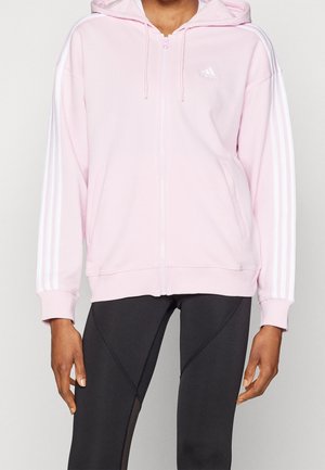 Personne portant un sweat à capuche zippé Adidas rose clair avec des bandes blanches sur les manches et un legging noir avec des coutures à motifs.