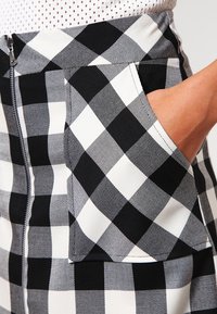 Sukně s černobílým kostkovaným vzorem, která má design postranní kapsy, je vyrobena z texturované látky a má viditelný zip. Detailní záběr na oblast kapsy.