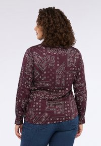 Blusa a maniche lunghe di color bordeaux con motivi paisley e floreali bianchi. Tessuto morbido, design aderente e orlo arrotondato. Abbinata a jeans blu.